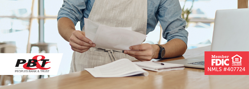 Business owner with apron on reviewing paperwork at desk in front of laptop at his business